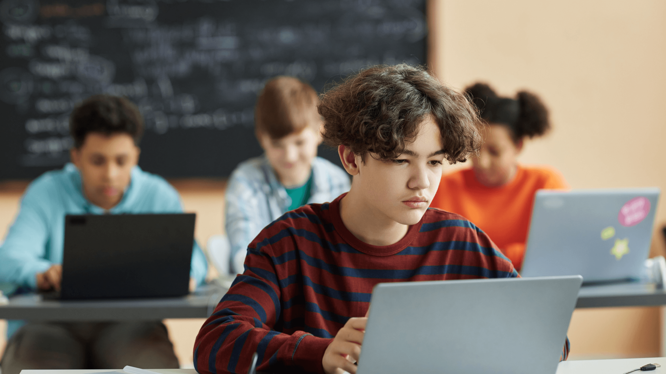 Image of a young student with brown hair and a burgundy and navy striped jumper using a laptop in a busy classroom