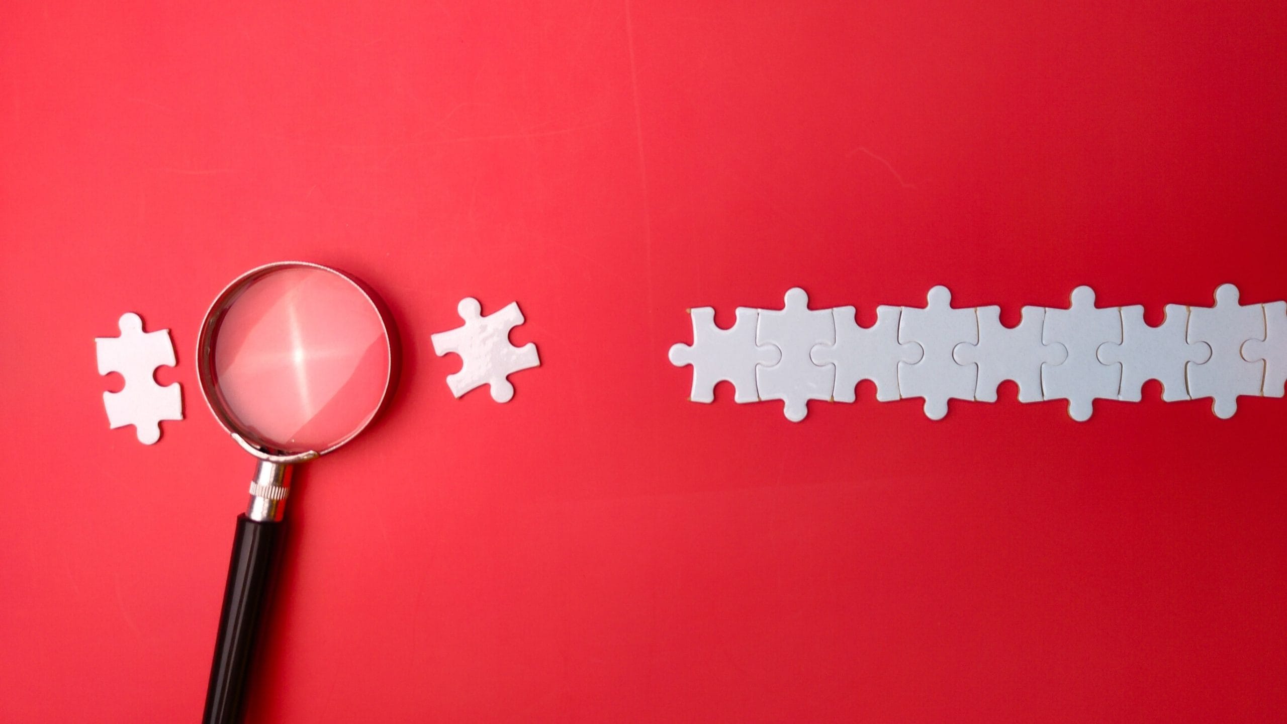 Red background with white jigsaw pieces and a magnifying glass missing one of the pieces