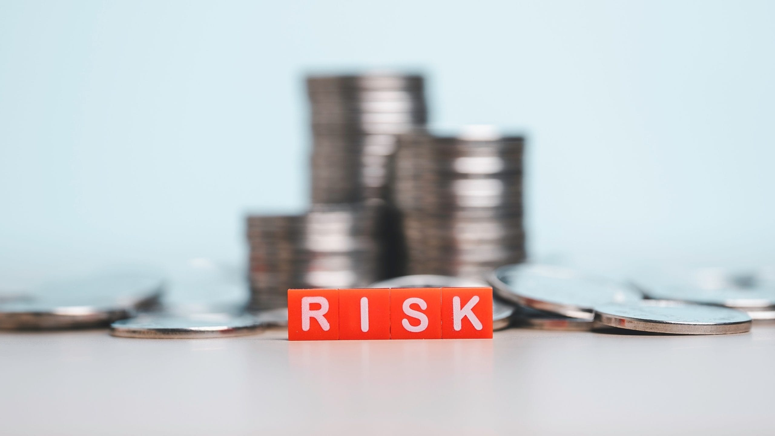Image of the word "risk" in red blocks in focus against a light blue background with stacks of coins